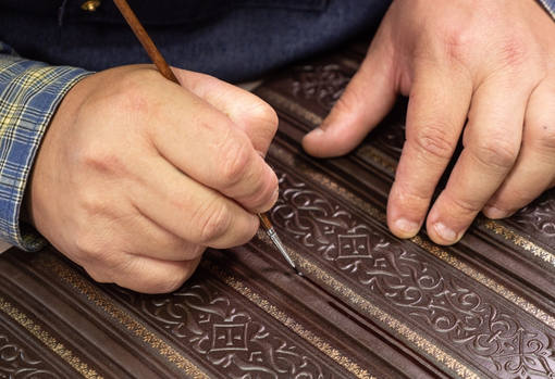 conservation-small-01 Mans hands shown holding a fine paintbrush as he restores some intricate detailing on an antique wooden item.